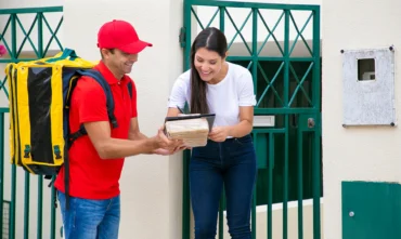 cheerful-brunette-woman-signing-delivery-sheet-with-pen-smiling-postman-with-thermo-backpack-holding-clipboard-parcel-standing-wearing-red-uniform-delivery-service-post-concept